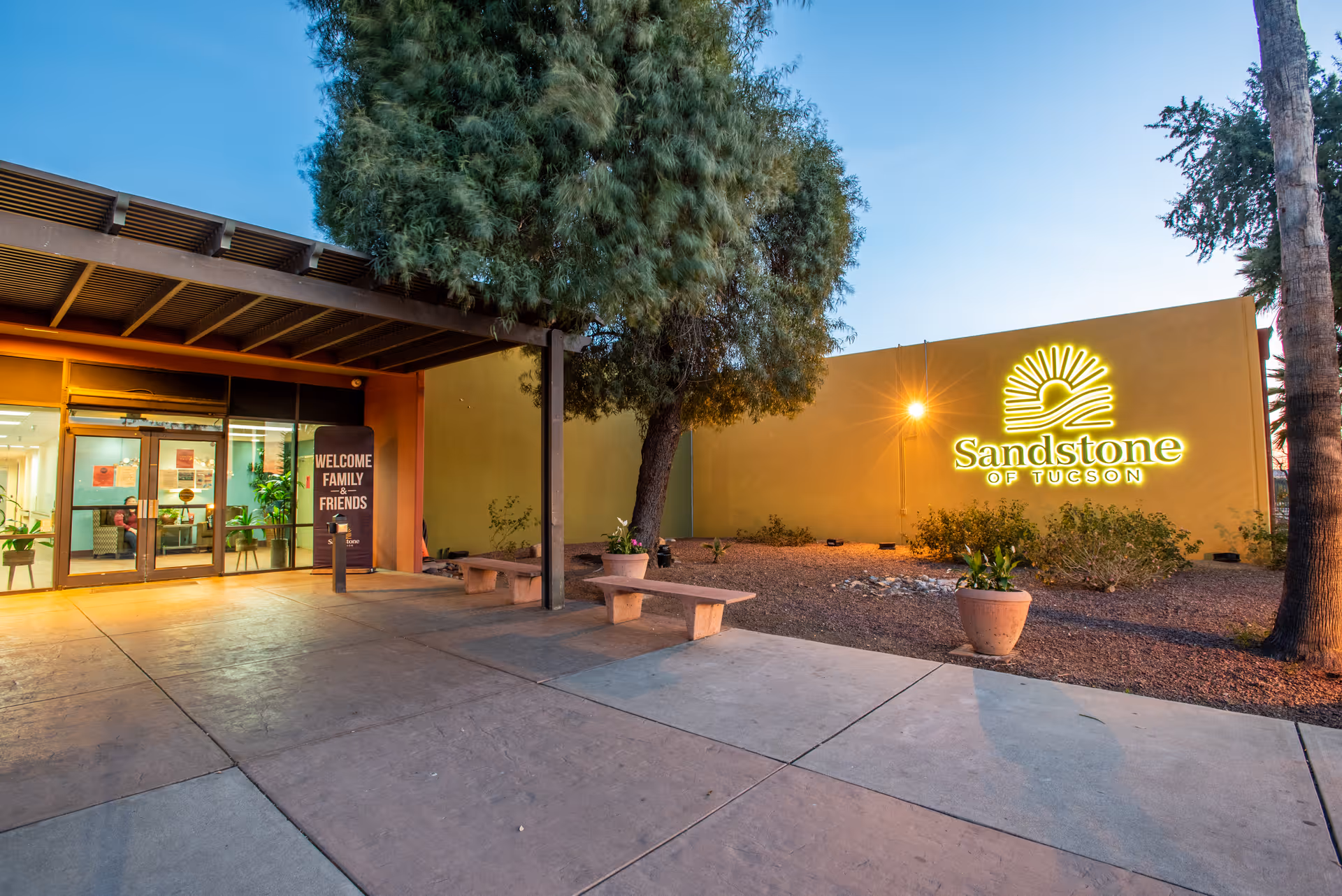 Exterior view of the entrance to Sandstone of Tucson facility at dusk, showing a covered walkway, benches, potted plants, a large tree, and a brightly lit sign with the facility's name on a yellow wall.