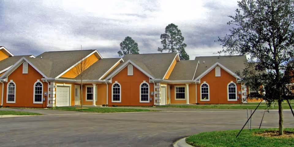 Exterior view of a single-story residential building with orange and beige walls, multiple gabled roofs, and several windows. There is a paved driveway and some grass with a tree in the foreground under a cloudy sky.