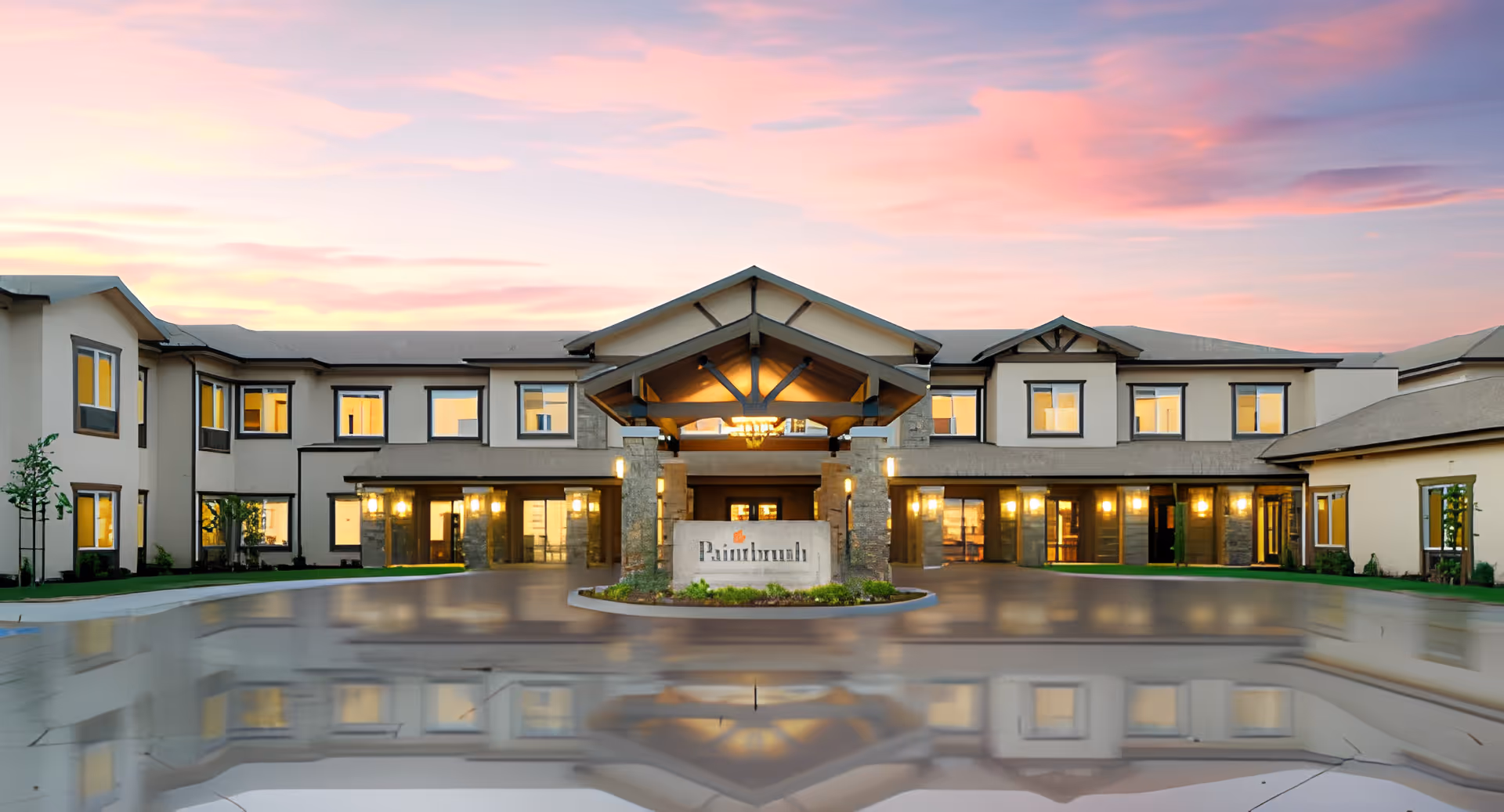 Front entrance of the Paintbrush Assisted Living building at sunset, showing a two-story facade with a canopy, lit windows, and reflections on wet pavement.
