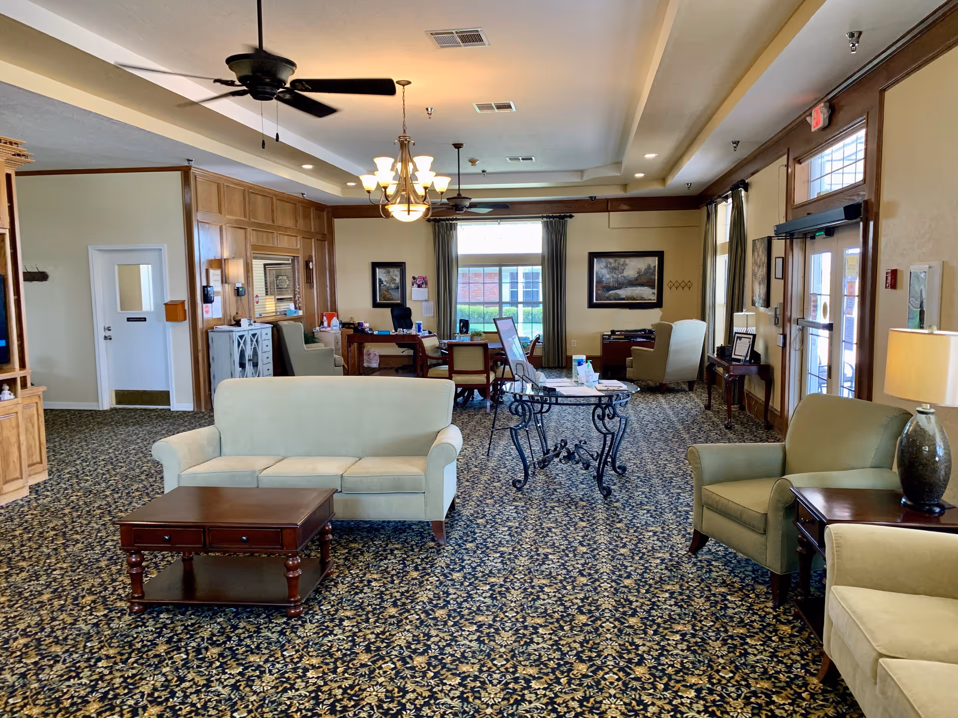 A spacious senior living facility common area with patterned carpet, beige sofas and armchairs, wooden coffee tables, ceiling fans, and a chandelier. The room has large windows with curtains, framed artwork on the walls, and a glass door entrance on the right side. There is a desk with chairs and office supplies in the background.