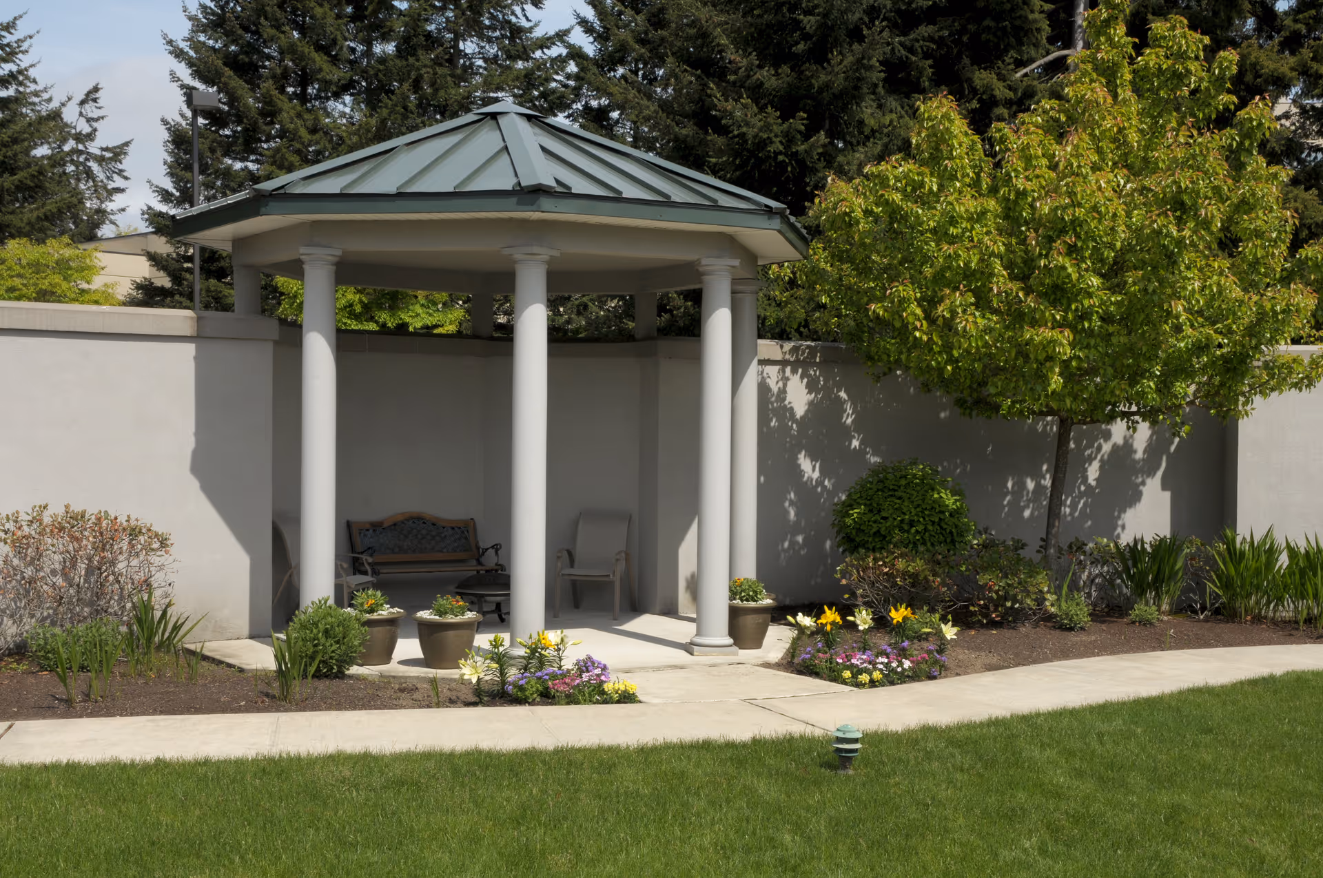 A small outdoor gazebo with a green metal roof and white columns, situated on a concrete patio surrounded by a garden with various plants, flowers, and a tree. There is a bench and a chair inside the gazebo. The area is enclosed by a light-colored wall with tall trees visible in the background.