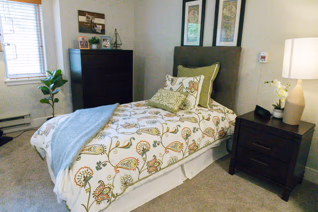 A neatly made single bed with floral patterned bedding and multiple pillows in a cozy bedroom. Next to the bed is a dark wooden nightstand with a lamp, a small plant, and a decorative item. A dark wooden dresser with framed photos and decor is against the wall near a window with blinds. The room has beige carpet and light-colored walls with two framed pictures above the bed.
