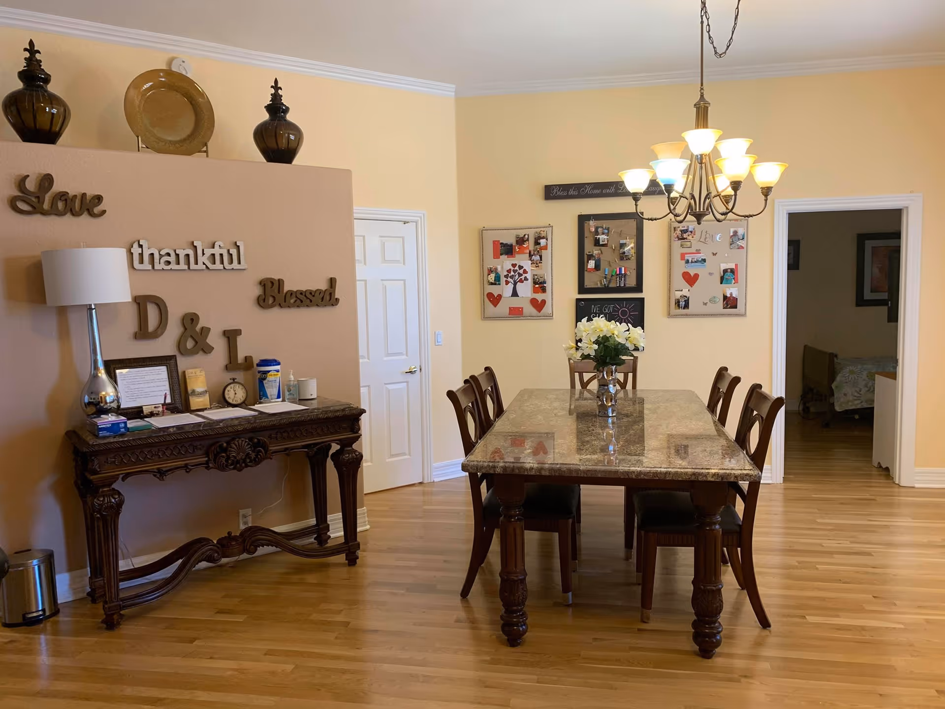 A dining room with a rectangular marble-top table surrounded by six wooden chairs. A chandelier with multiple lights hangs above the table. On the left wall, there is a decorative console table with a lamp, clock, and other items. Above the console table, words like 'Love', 'thankful', 'Blessed', and 'D & L' are mounted on the wall. The back wall has three bulletin boards with photos and decorations. Two doorways are visible, one leading to another room with a bed.