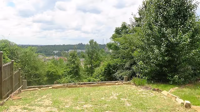 Backyard lawn with a low stone border, a wooden fence at left, and trees overlooking a distant landscape under a cloudy sky.