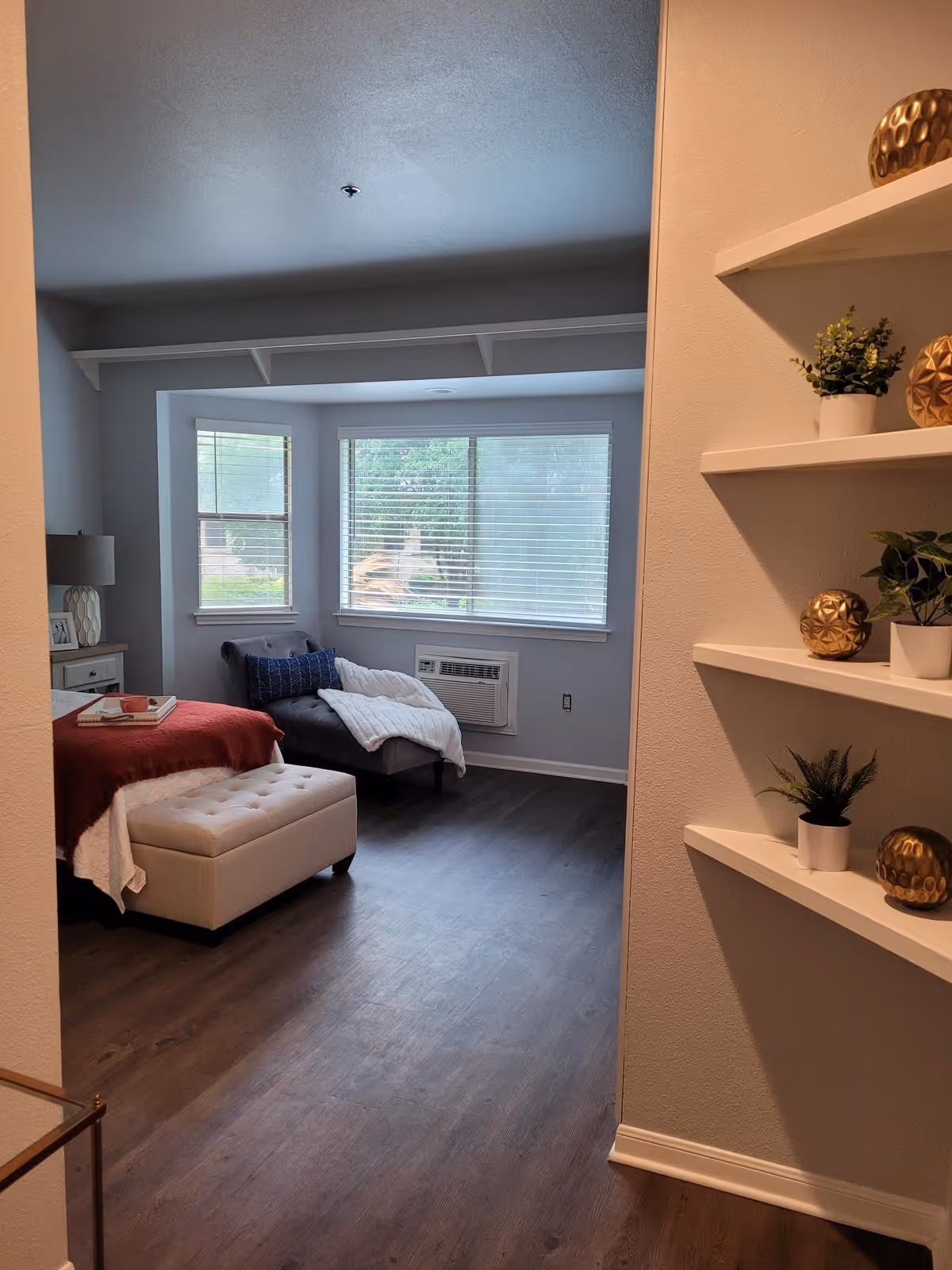 View into a bedroom with a large window featuring horizontal blinds. The room has a chaise lounge with a white blanket and a blue pillow, a bed with a red blanket, and a white ottoman at the foot of the bed. To the right, there are white wall shelves decorated with small potted plants and gold decorative spheres. The floor is a dark wood laminate.