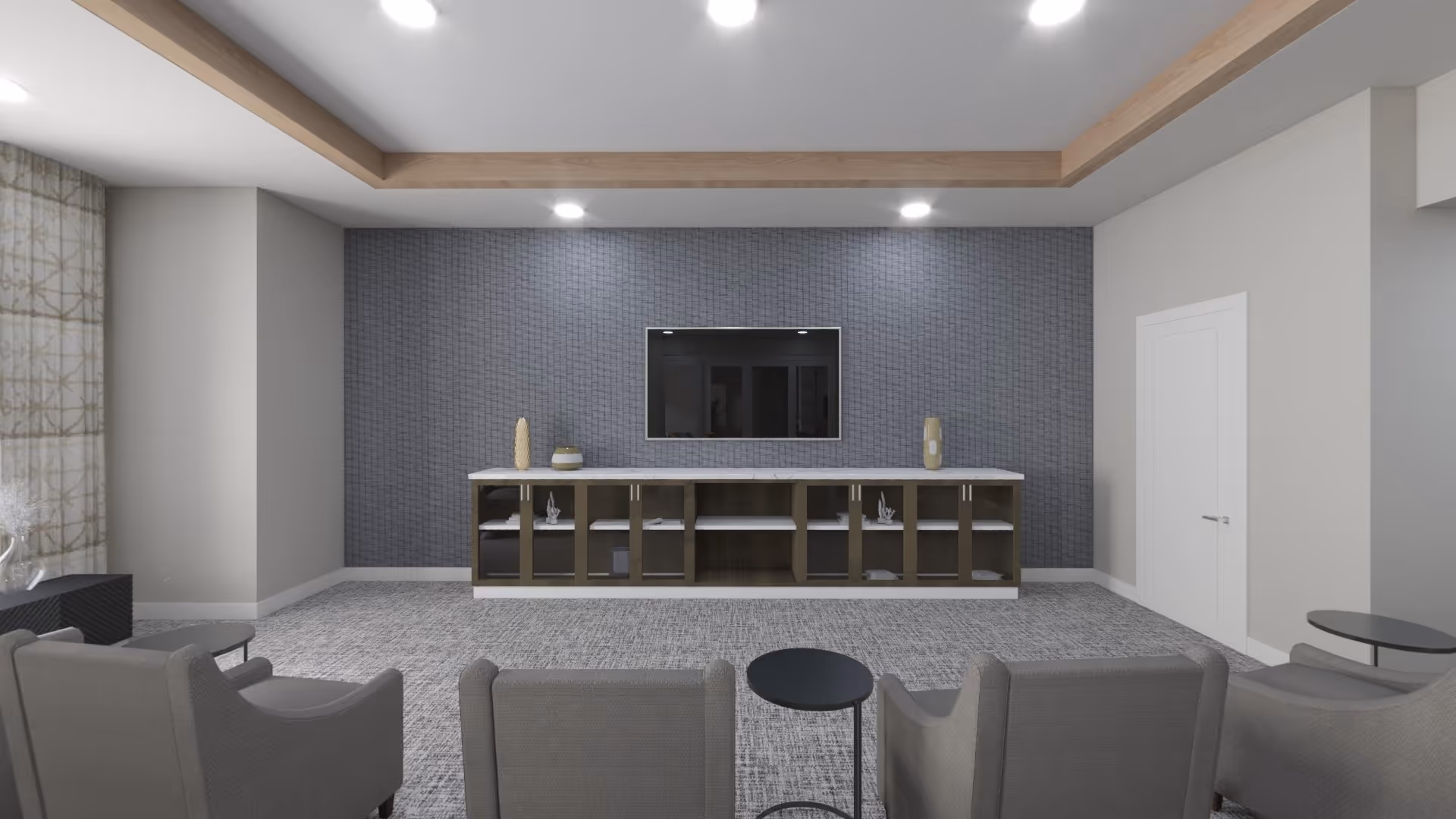 A modern living room with a gray patterned carpet, four gray armchairs arranged in two rows facing a wall-mounted flat screen TV. Below the TV is a long wooden cabinet with glass doors and decorative items on top. The walls are painted light gray with one accent wall behind the TV featuring a textured gray pattern. The ceiling has recessed lighting and a wooden trim detail.