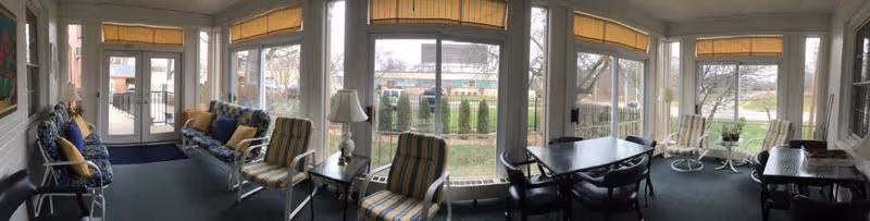 Panoramic sunroom with armchairs, sofas, side tables and a dining table facing large windows overlooking a lawn.