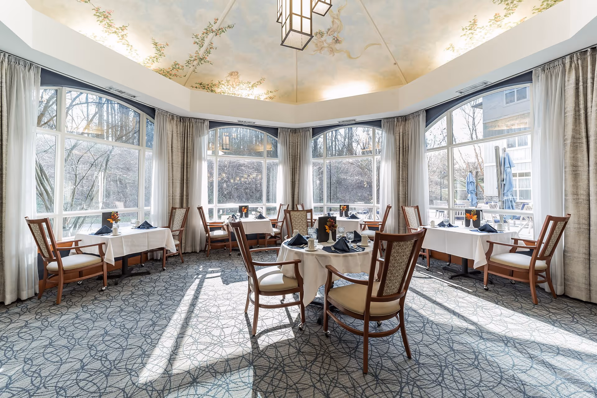 Bright dining room with large windows letting in natural light, round and rectangular tables set with white tablecloths, napkins, and small floral centerpieces, wooden chairs with cushioned seats, and a decorative ceiling with floral artwork.