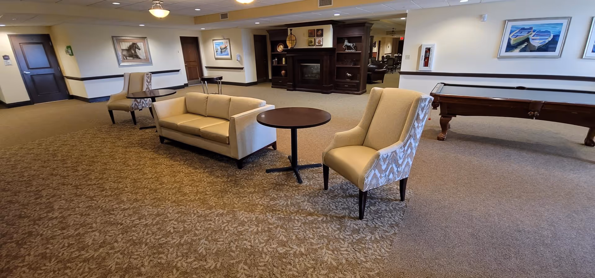 Spacious senior living common area with sofas and armchairs around a round table, a fireplace cabinet and a pool table in the background.