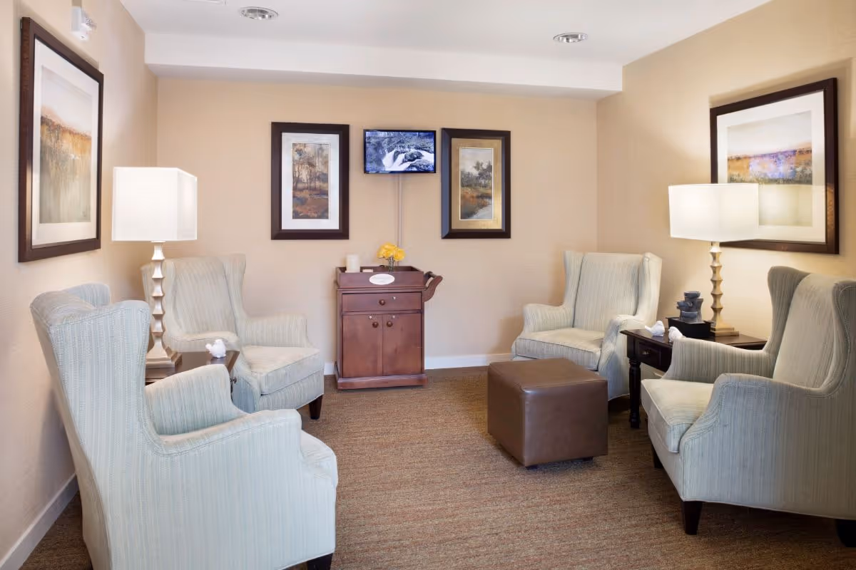 Cozy seating area with four upholstered armchairs arranged around a leather ottoman, side tables with lamps, framed artwork, and a small cabinet under a wall-mounted TV.