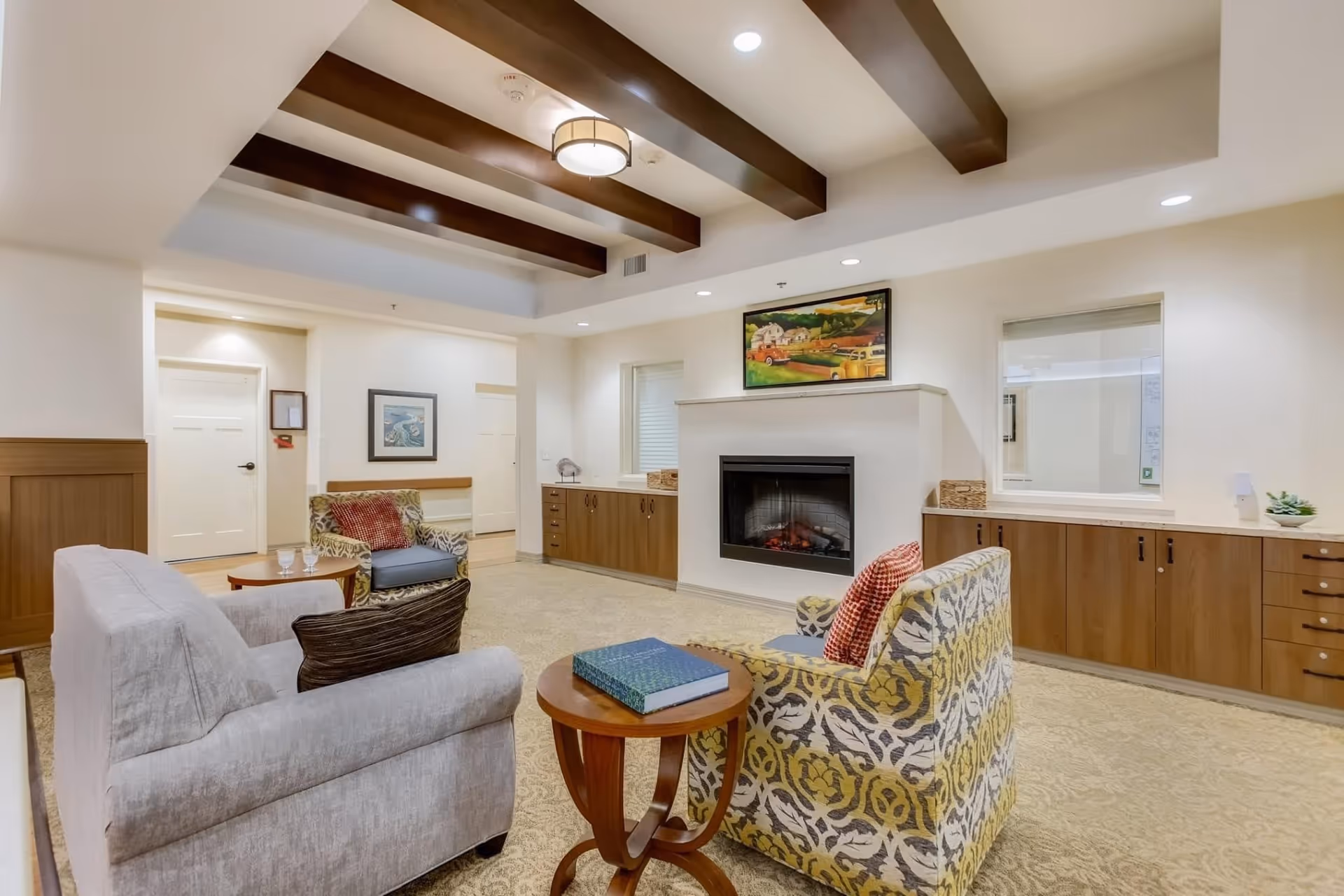 A cozy living room area with a beige carpet, two patterned armchairs with red pillows, a light gray sofa with a dark pillow, and a small wooden side table with a book on it. The room features a white fireplace with a colorful painting above it, wooden cabinets along the walls, and exposed dark wooden ceiling beams with a round ceiling light fixture.