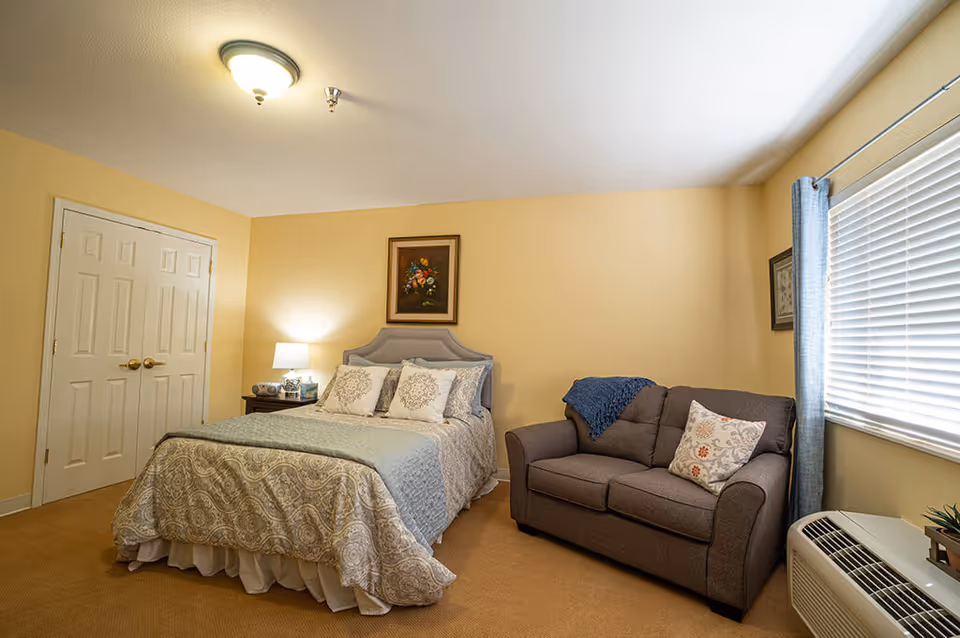 A cozy bedroom with a made bed, nightstand and lamp, a loveseat by a window with blinds, and neutral decor.