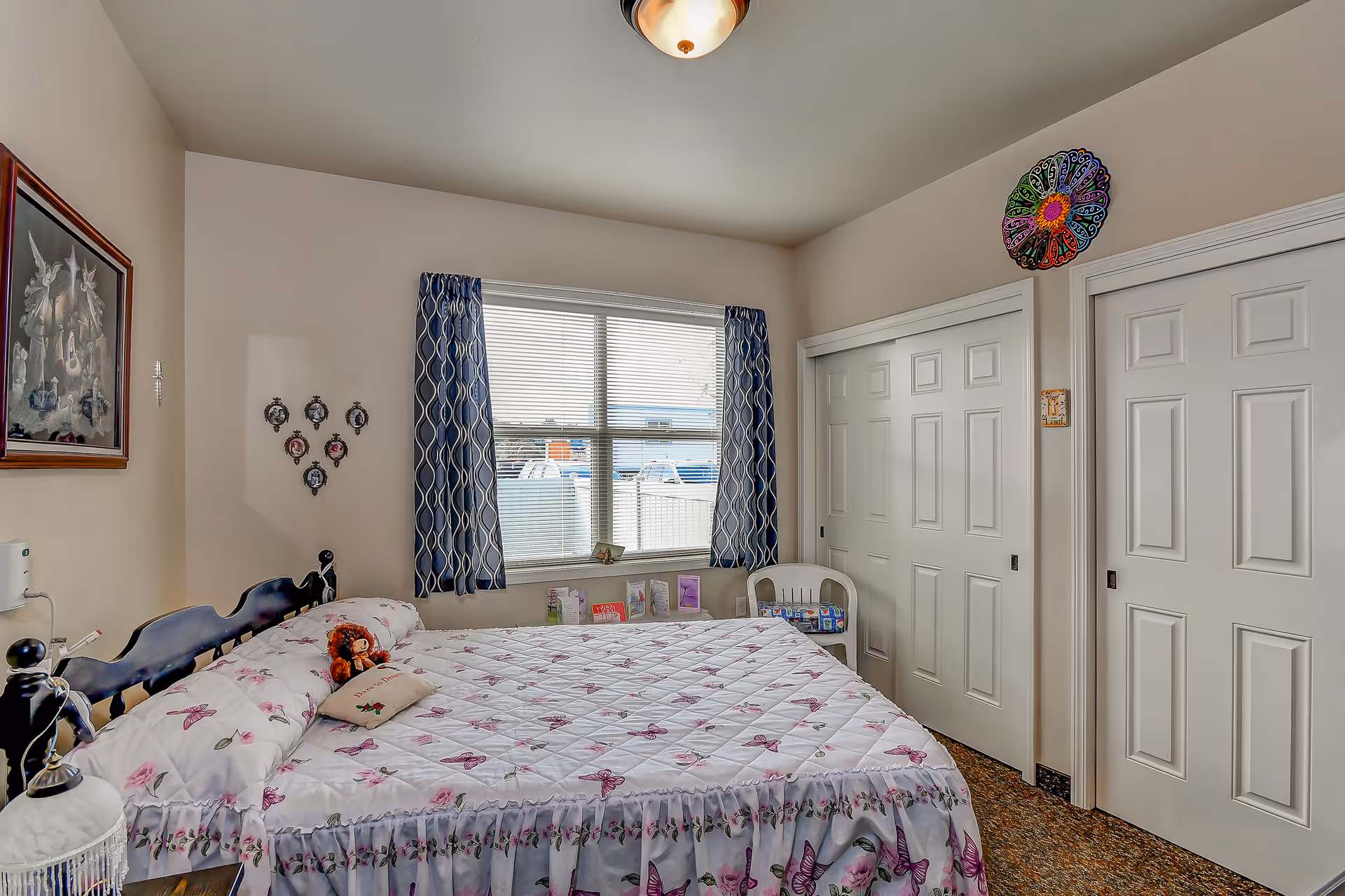 A cozy bedroom with a bed covered in a white quilt with purple butterfly patterns. There is a small teddy bear and a decorative pillow on the bed. The room has a window with blue patterned curtains, two white sliding closet doors, a white chair, and a framed picture on the wall. The walls are beige, and the floor has a patterned carpet.