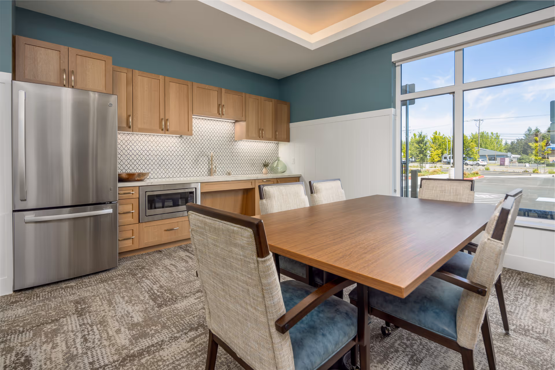 Sunlit dining area with a wooden table and upholstered chairs next to a small kitchen with a stainless steel refrigerator and wood cabinets.