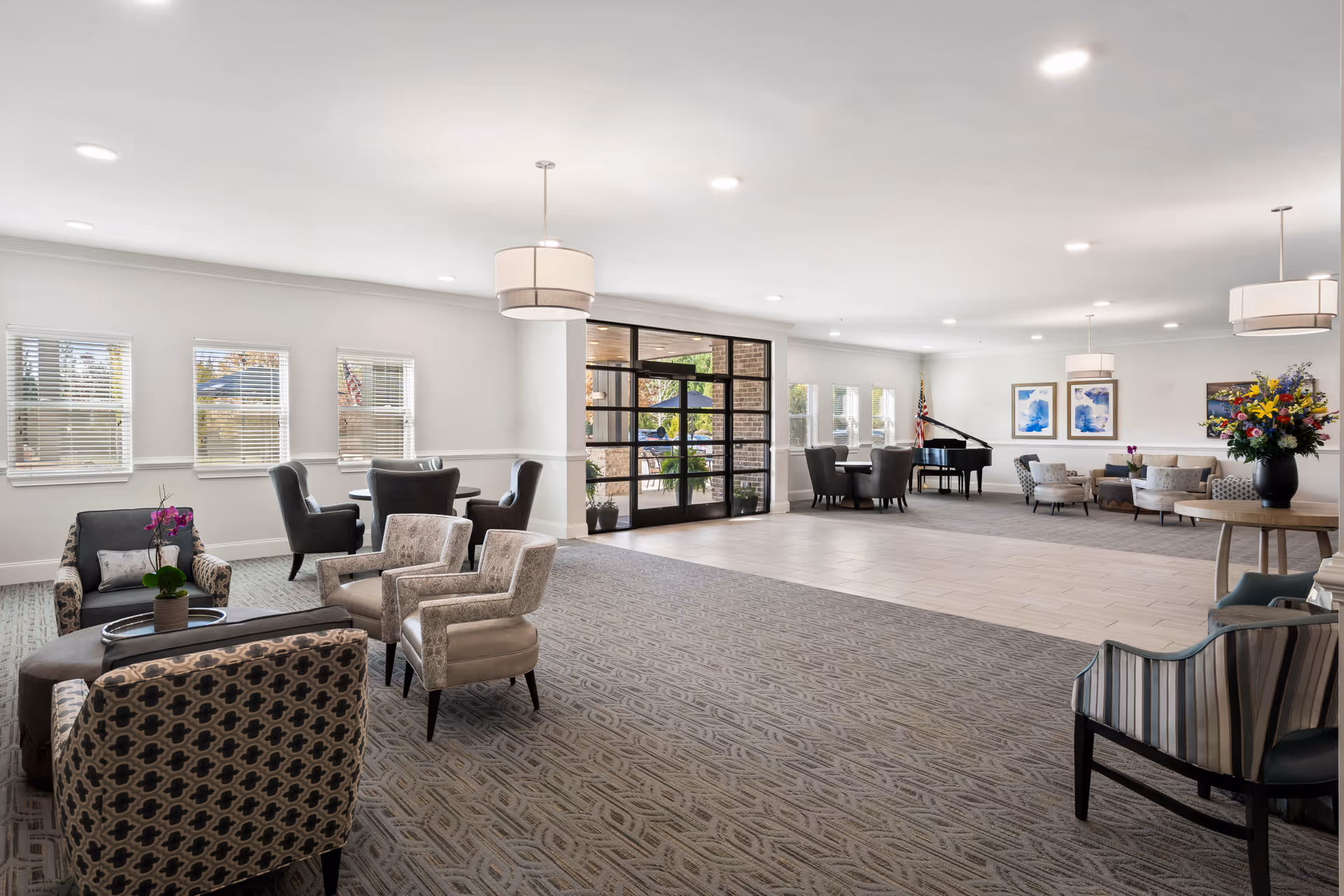 A spacious, well-lit common area in The Mansions at Alpharetta Independent Living featuring multiple seating arrangements with upholstered chairs and small tables. The room has large windows with blinds, a glass door leading outside, a grand piano in the background, and decorative artwork on the walls. The floor is carpeted with a patterned design, and there are modern pendant lights hanging from the ceiling. A vase with colorful flowers is placed on a round table to the right.