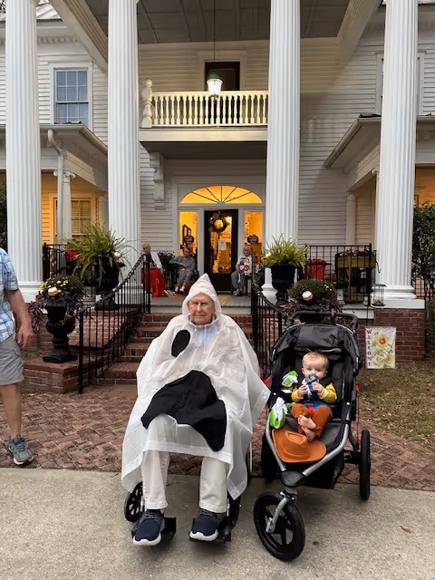 An elderly person in a wheelchair wearing a white poncho with a black spot, sitting next to a baby in a stroller. They are outside a large white building with tall columns and a porch decorated with plants and a wreath on the door. A person in shorts and sneakers is partially visible on the left side of the image.