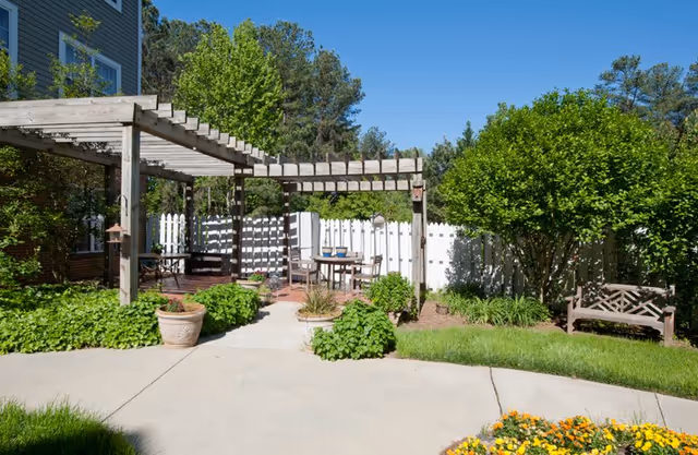 Outdoor patio area with a wooden pergola, several chairs and tables, surrounded by green bushes, trees, and a white picket fence under a clear blue sky.