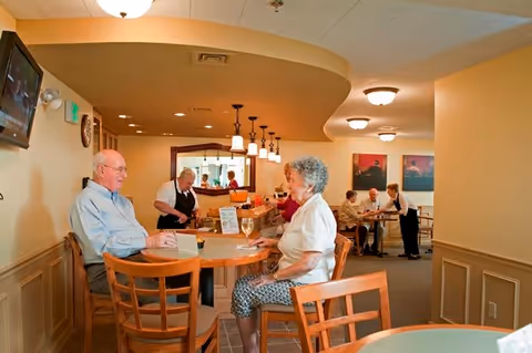 An elderly man and woman sitting at a round wooden table in a dining area, with a waiter serving drinks. In the background, other elderly people are seated at tables, and a staff member is attending to them. The room has warm lighting and beige walls.