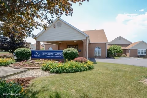 Exterior view of Keystone Woods senior living community building with a covered entrance, surrounded by green lawn, bushes, and trees under a clear blue sky.