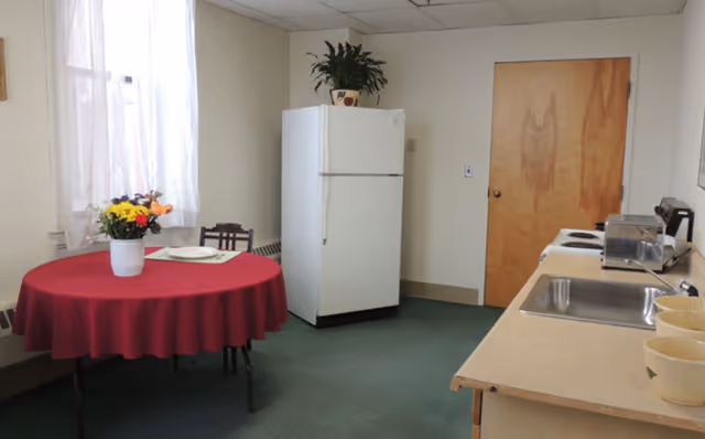 Small kitchen and dining area with a round table covered by a red tablecloth, a white refrigerator, countertop with sink and appliances, and a closed wooden door.