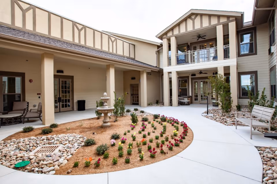 Outdoor courtyard area at The Villages of Windcrest featuring a small garden with colorful flowers, a stone water fountain, benches, and a two-story building with covered patios and ceiling fans.