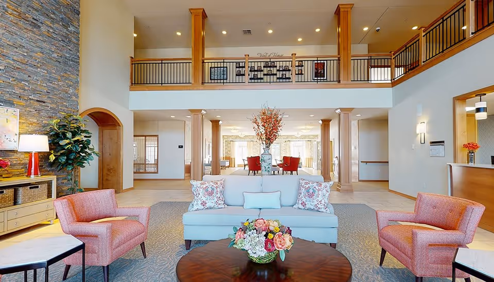 A spacious and well-lit senior living facility common area with a high ceiling and a balcony railing above. The room features a light blue sofa with floral pillows, two pink armchairs, a round wooden coffee table with a floral arrangement, and a stone accent wall with a lamp and plant. There is a reception desk on the right and a seating area with red chairs in the background.