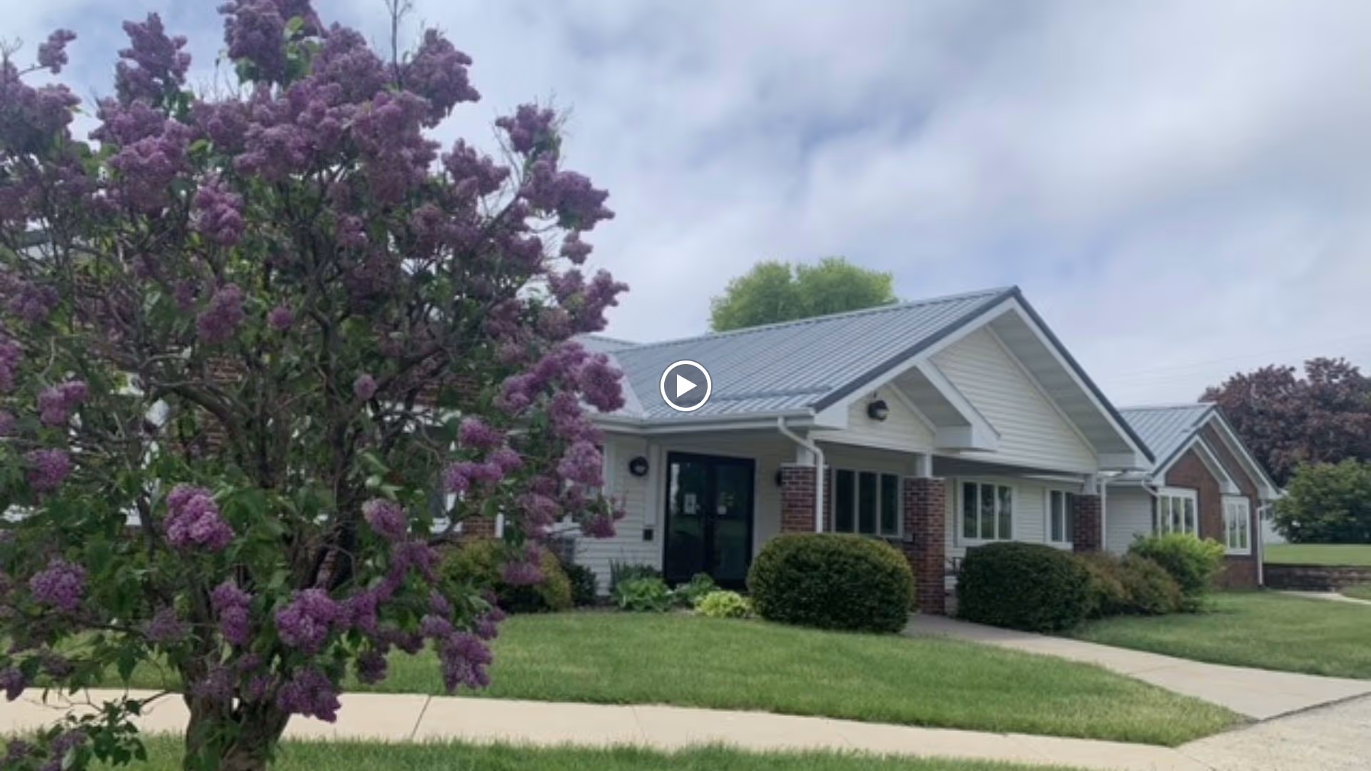 Single-story convalescent home building with a metal roof, front entrance, landscaped lawn and a large purple-flowering shrub.