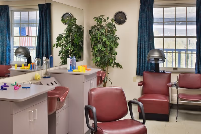 Interior of a hair salon area in a senior living facility with maroon salon chairs, a hair dryer, a large mirror, a potted plant, and windows with blue curtains. Various hair care products and colorful hair rollers are placed on the counter.