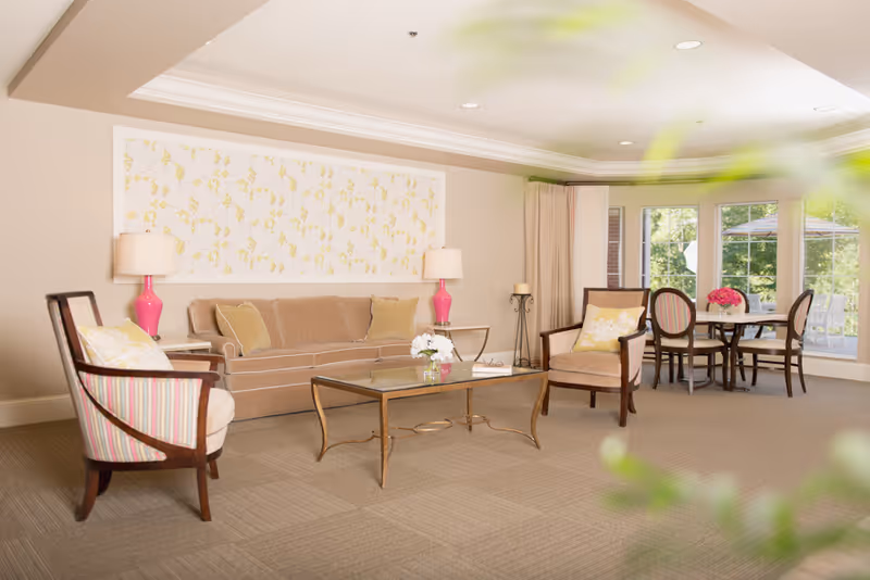 A bright and spacious living room area with a beige sofa, two armchairs with patterned cushions, a glass coffee table with flowers, and two pink table lamps on side tables. In the background, there is a round dining table with four chairs near large windows showing an outdoor patio with umbrellas.