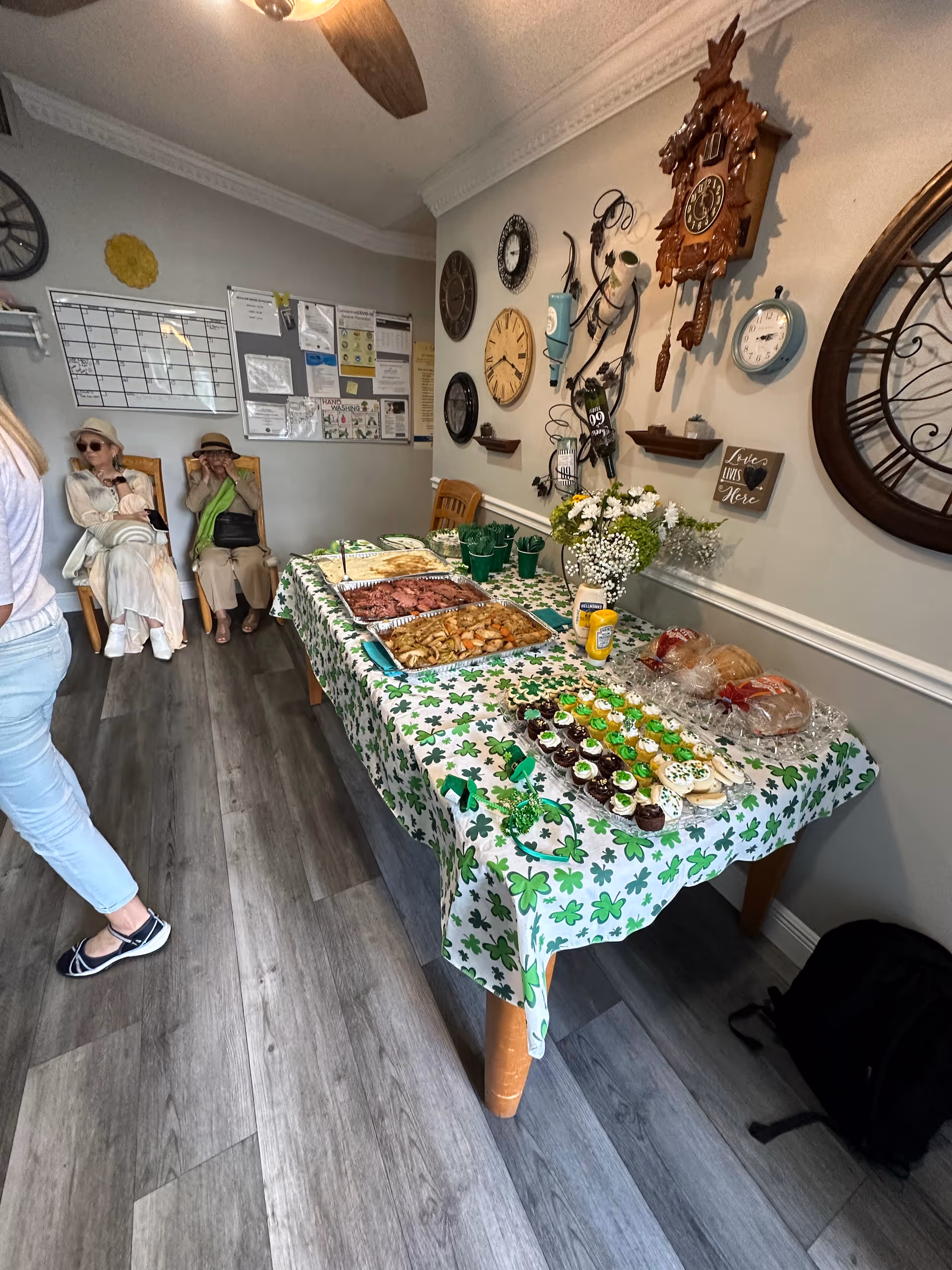 A St. Patrick's-themed buffet table with trays of food and cupcakes in a communal room decorated with wall clocks, while two seated people rest against the far wall.