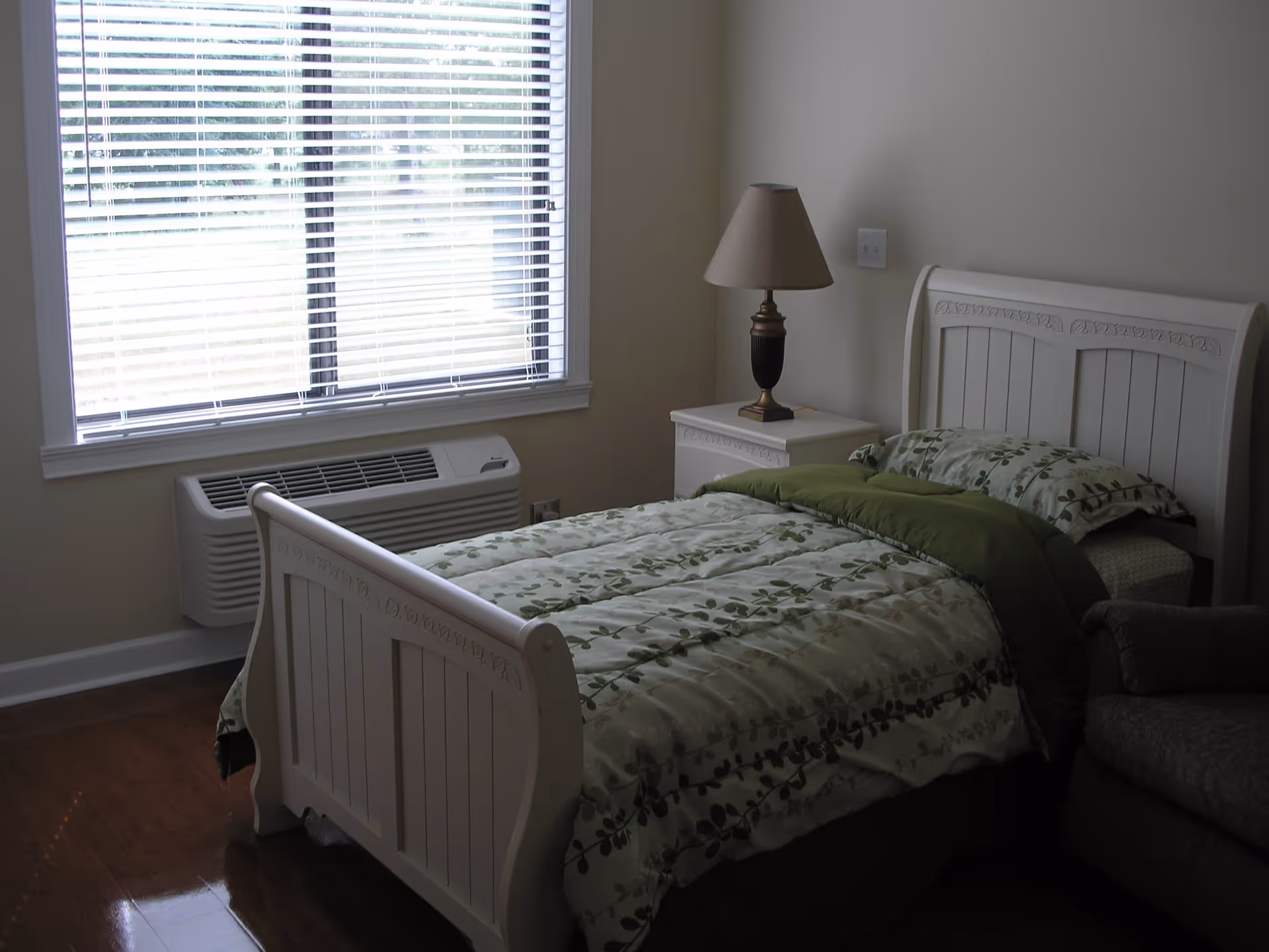 A bedroom with a white wooden bed frame and a green and white floral patterned comforter. There is a white nightstand beside the bed with a lamp on it. A window with white blinds is on the left wall, and a heating/cooling unit is installed below the window. The floor is wooden, and part of a gray upholstered chair is visible on the right side.
