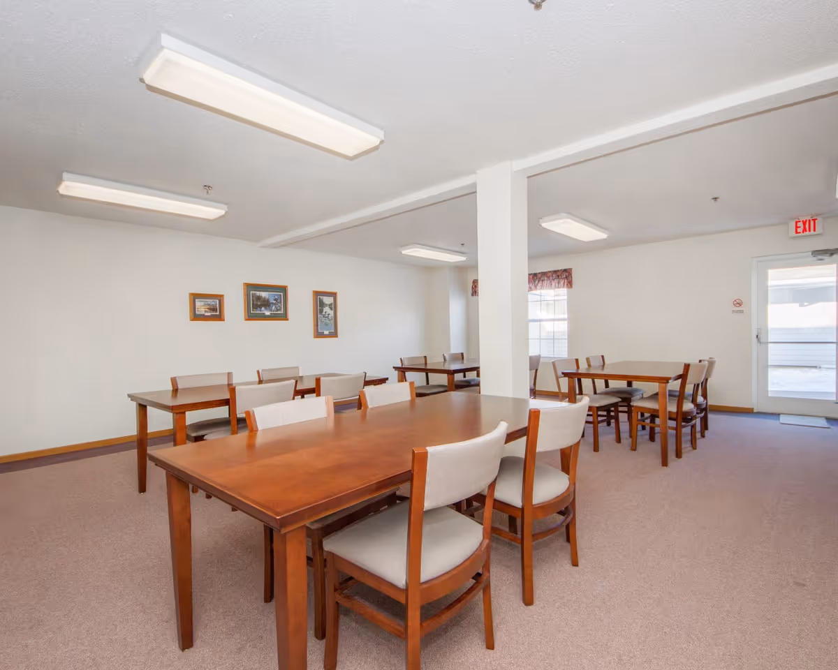 A dining room with multiple wooden tables and cushioned chairs arranged neatly. The room has white walls with three framed pictures, carpeted floor, fluorescent ceiling lights, a window with a valance, and an exit door with a red exit sign above it.