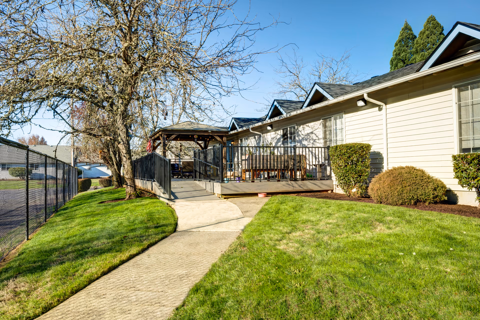 Outdoor view of a senior living facility with a paved walkway leading to a wooden deck area with a gazebo. The building is light-colored with several windows and surrounded by green grass, bushes, and leafless trees under a clear blue sky.