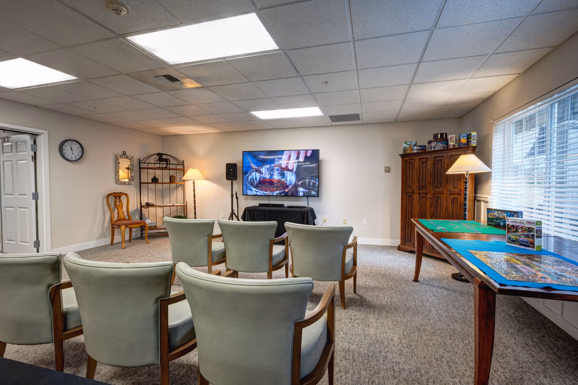 A cozy common room with several light green cushioned chairs arranged in rows facing a wall-mounted flat screen TV. To the right, there is a wooden table with puzzles and a floor lamp beside it. On the left side, there is a wooden chair, a small shelving unit, and a wall clock. The room has carpeted flooring, a drop ceiling with fluorescent lights, and a window with blinds on the right wall.