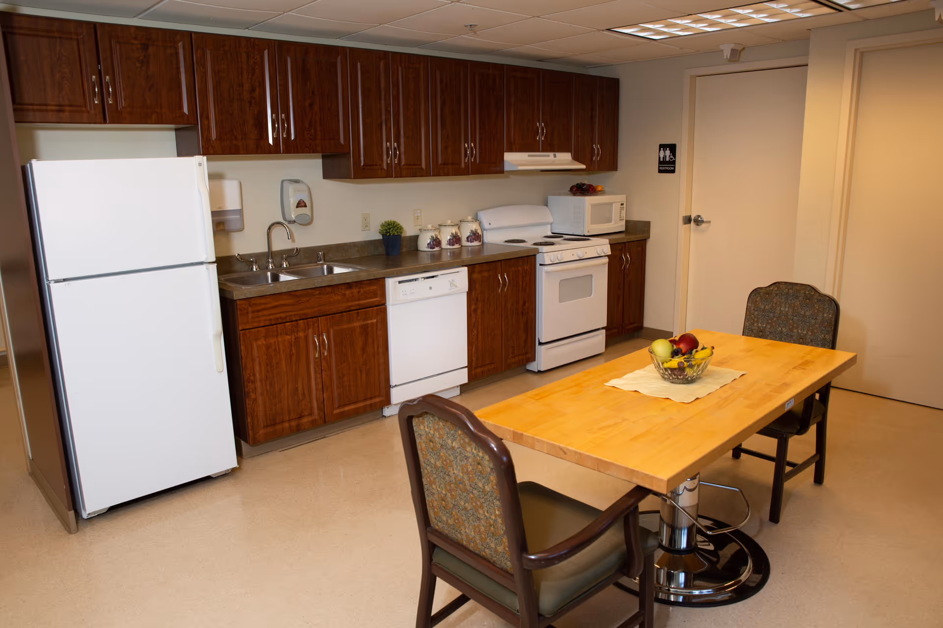 Small communal kitchen with wooden cabinets, white appliances, a sink, and a wooden table with chairs and a bowl of fruit.