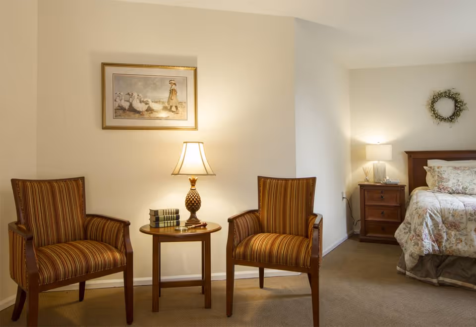 Cozy bedroom seating area with two striped armchairs and a round table with a lamp; a bed and nightstand with a lamp are visible on the right.