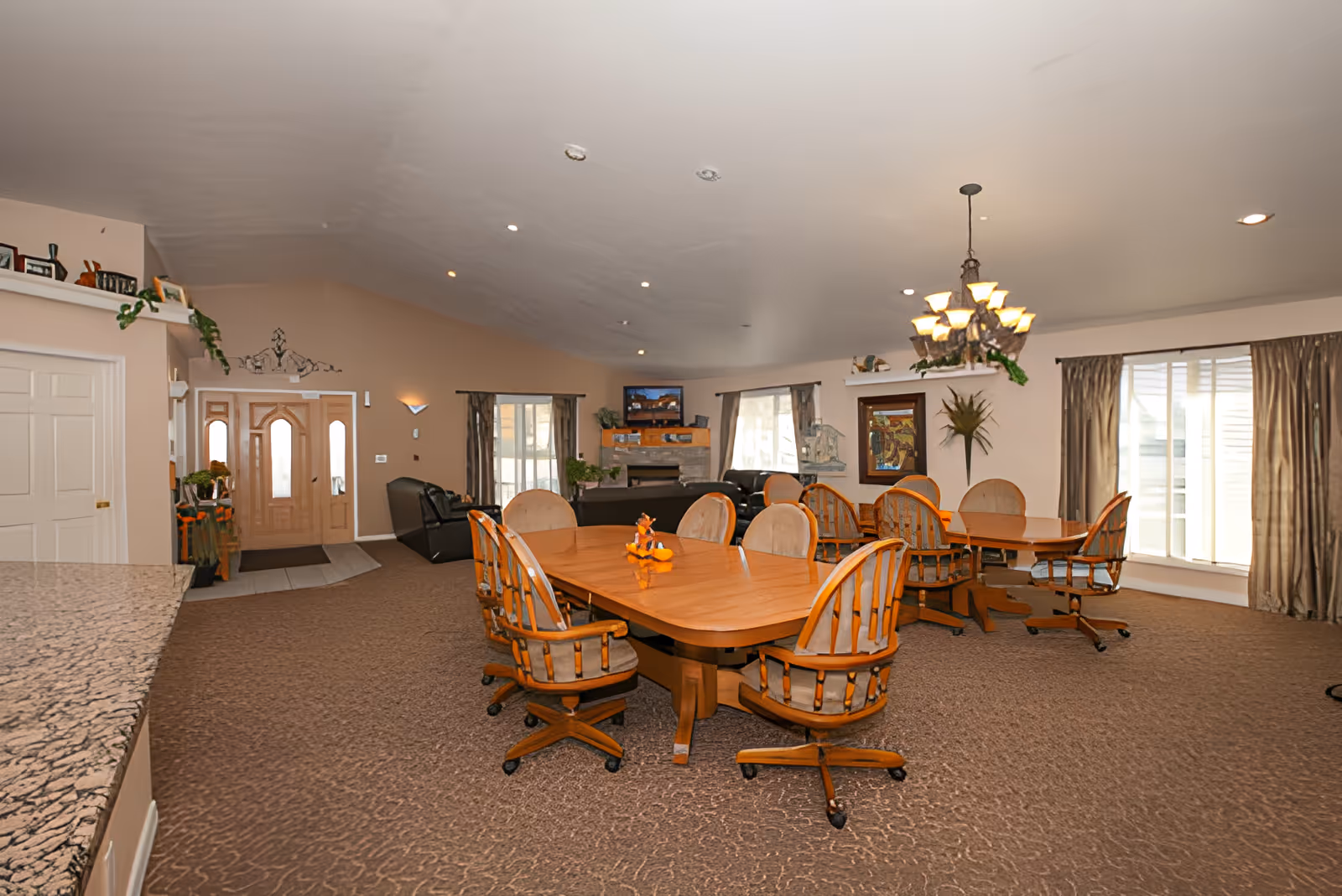 Spacious common area with two wooden dining tables surrounded by cushioned chairs on wheels, a chandelier hanging from the ceiling, large windows with curtains, a fireplace with a TV above it, and a seating area with black leather chairs near the entrance door.