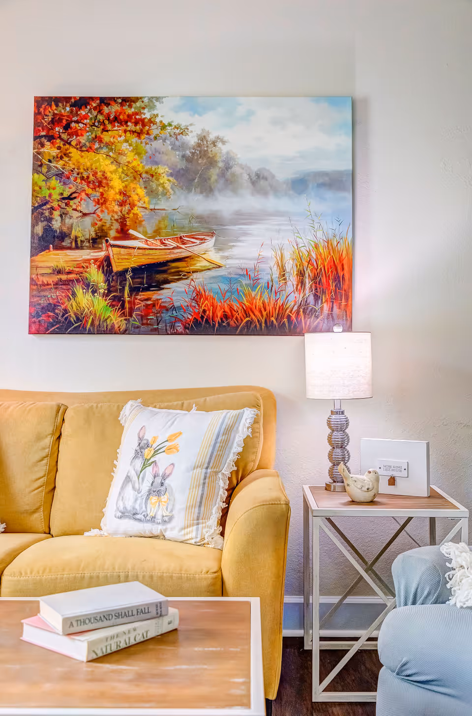 A cozy living room area featuring a mustard yellow couch with a decorative pillow showing rabbits and flowers. Above the couch hangs a colorful painting of a lakeside scene with boats and autumn foliage. To the right of the couch is a small side table with a lamp, a bird figurine, and a framed sign. In the foreground, a wooden coffee table holds two books.