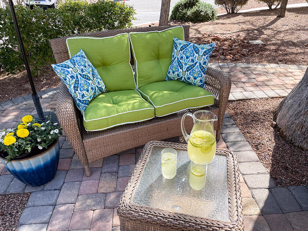 Outdoor patio area with a wicker loveseat featuring bright green cushions and two blue patterned throw pillows. In front of the loveseat is a matching wicker table with a glass top holding a pitcher of lemonade with lime slices and two glasses of lemonade. There is a blue flower pot with yellow and white flowers to the left, and the area is paved with multicolored bricks surrounded by landscaping with rocks, bushes, and trees.