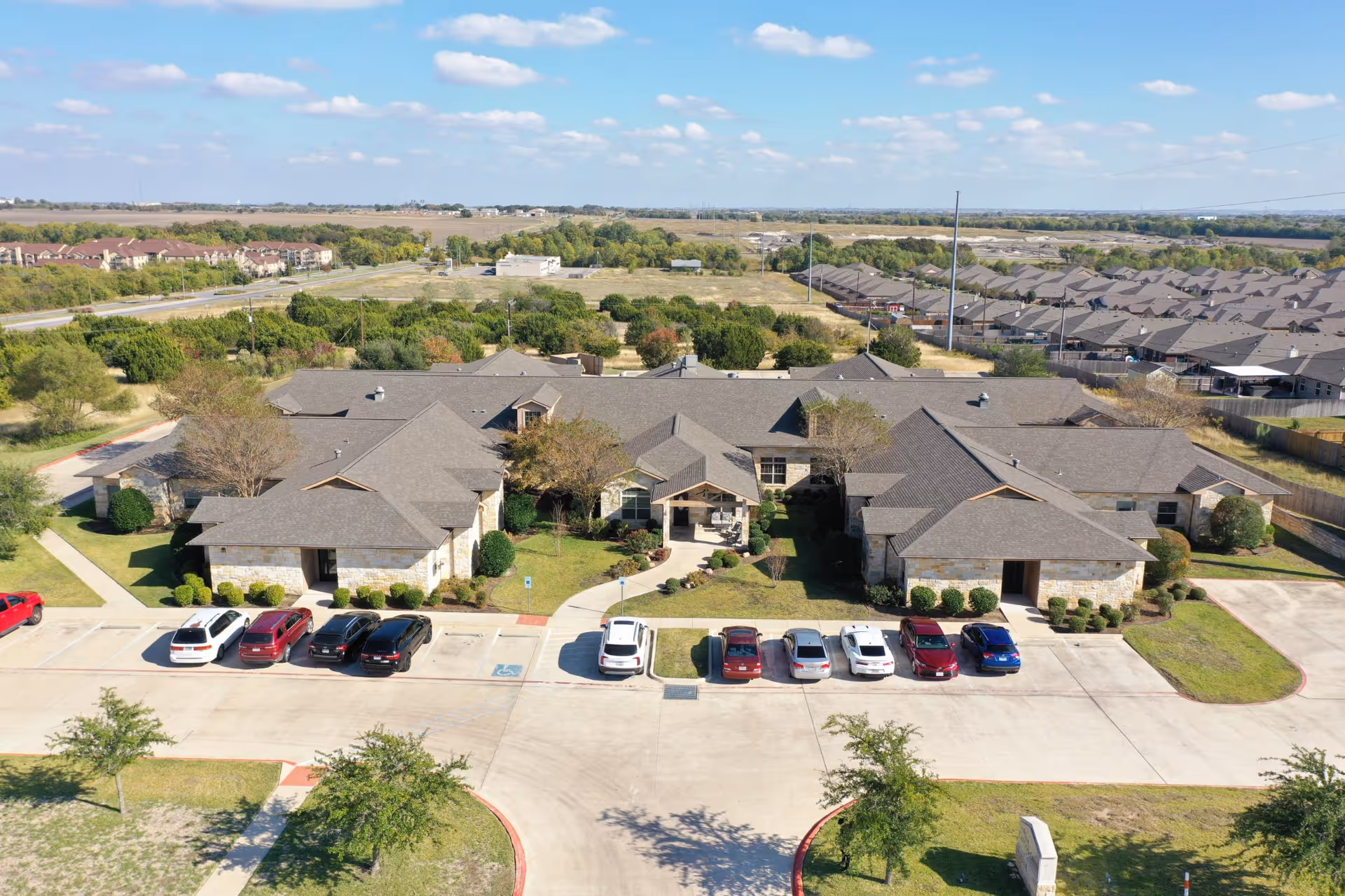 Aerial view of a single-story senior living facility named Canyon Creek Memory Care, surrounded by parking spaces with several cars parked, landscaped greenery, and neighboring residential areas under a partly cloudy sky.