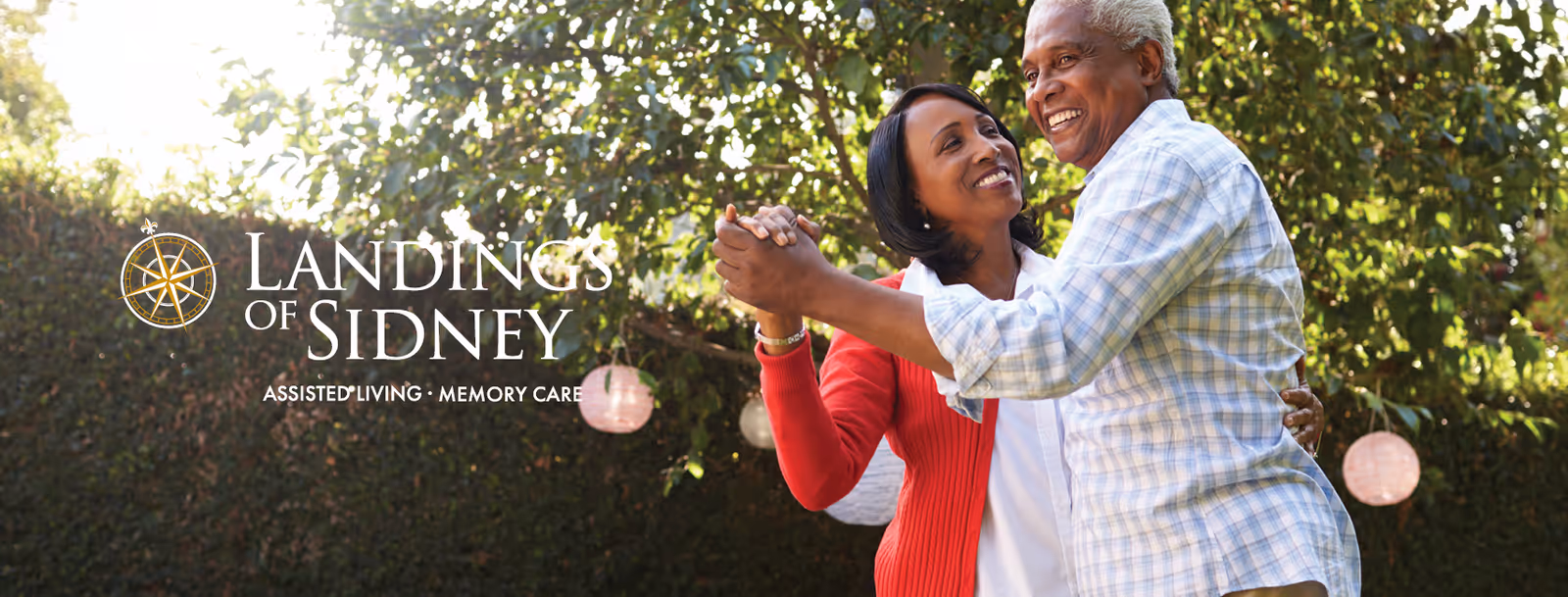 An elderly couple dancing outdoors in a garden area with greenery and hanging lanterns in the background. The woman is wearing a red cardigan and the man is wearing a light blue checkered shirt. The image includes the text 'Landings of Sidney Assisted Living · Memory Care' on the left side.