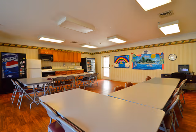 A communal dining area with several tables and chairs arranged in rows. The room has wooden flooring, a kitchenette with wooden cabinets, a refrigerator, microwave, stove, and a Pepsi vending machine. Colorful children's artwork is displayed on the wall, and a small TV is placed on a desk in the corner.