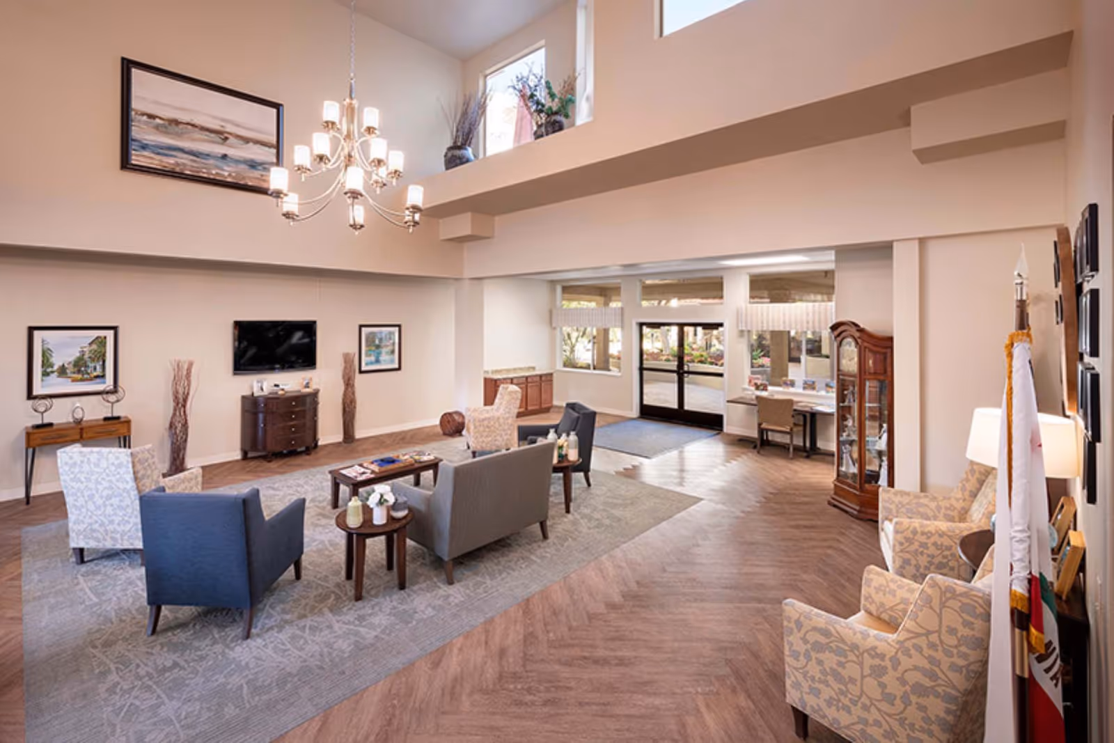 A spacious and well-lit living room area in Holiday Warburton Place featuring a chandelier, multiple armchairs and sofas arranged around a coffee table, a wall-mounted TV, framed artwork, and large windows near the entrance letting in natural light.