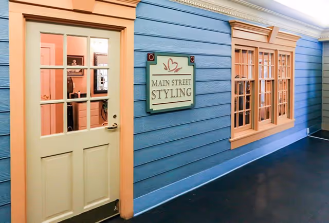 Interior hallway with blue siding walls and peach trim around a door and windows. The door has glass panes and is partially open, showing a room inside with a mirror and hair styling equipment. A sign on the wall reads 'Main Street Styling'.