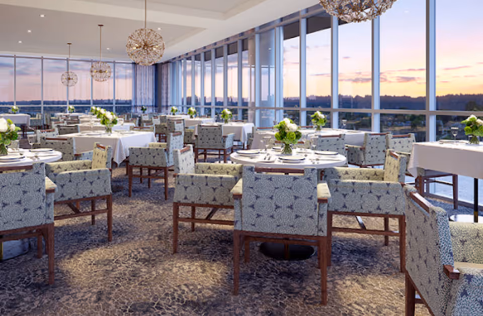 A spacious dining room with multiple tables covered in white tablecloths, each set with plates, glasses, and floral centerpieces. The room features large floor-to-ceiling windows offering a panoramic view of the sunset outside. Modern chandeliers hang from the ceiling, and patterned upholstered chairs surround the tables.