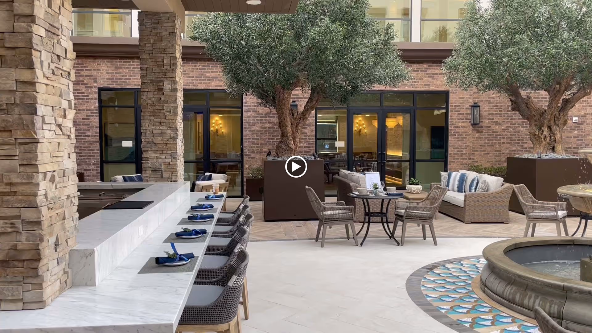 Outdoor courtyard area with a stone fountain in the foreground, surrounded by seating including a marble countertop with chairs, round tables with chairs, and cushioned sofas. There are large potted trees and a brick building with glass doors and windows in the background.