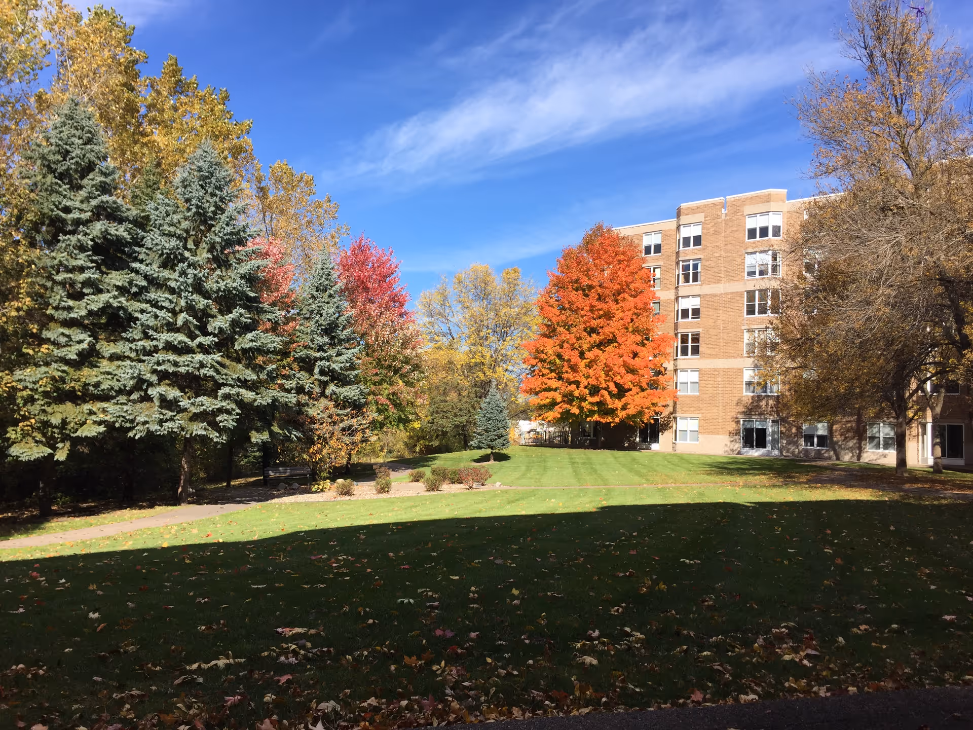 A senior living facility building with a brick exterior surrounded by a well-maintained lawn and various trees displaying autumn colors under a clear blue sky.