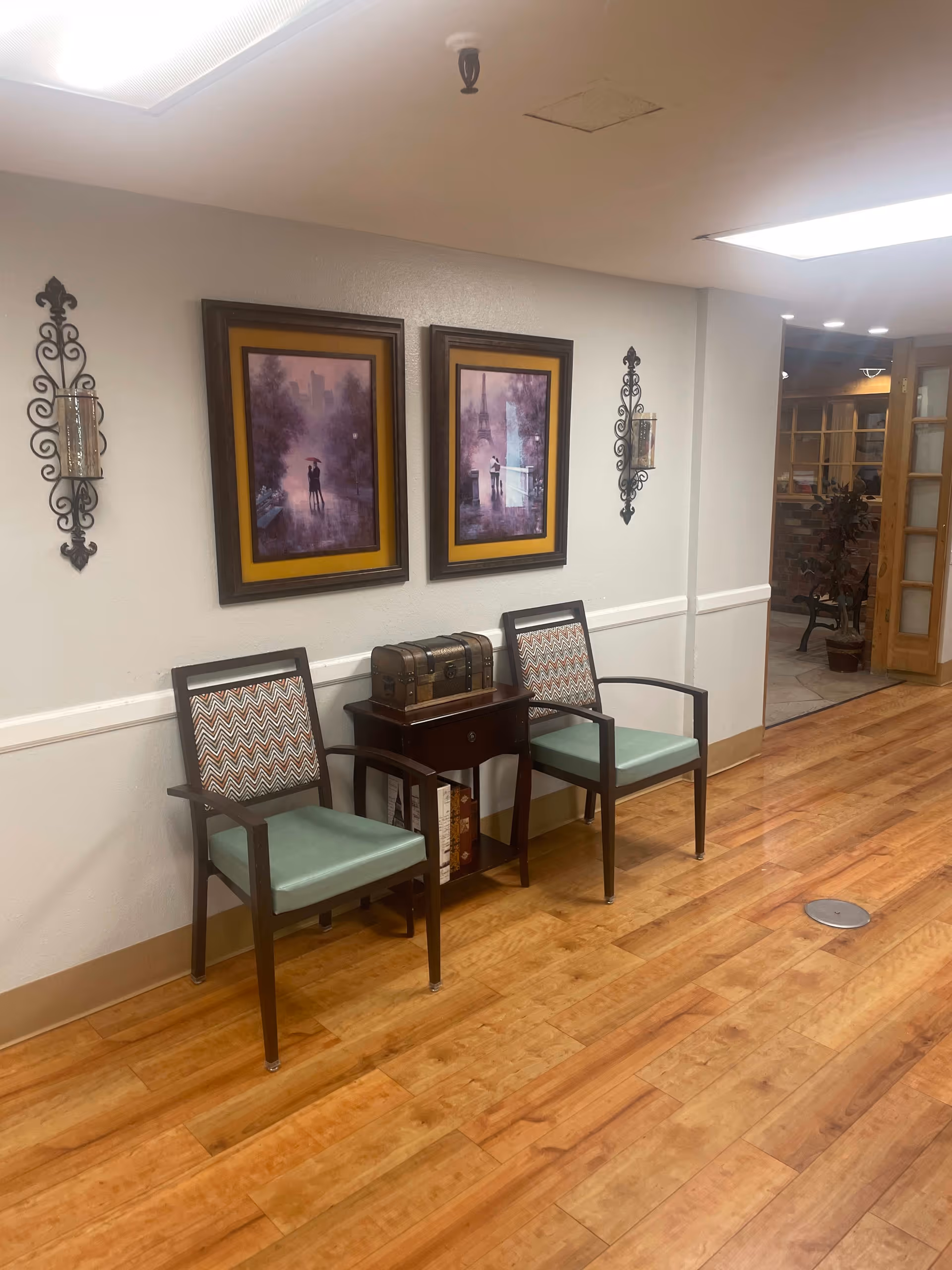 Hallway seating area with two chairs, a small side table holding a decorative chest, framed artwork on the wall and wood-look flooring.