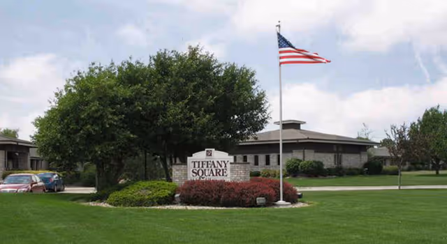 Exterior view of Tiffany Square facility with a large green lawn, a flagpole flying the American flag, a sign reading 'Tiffany Square' surrounded by bushes and trees, and a building in the background under a cloudy sky.