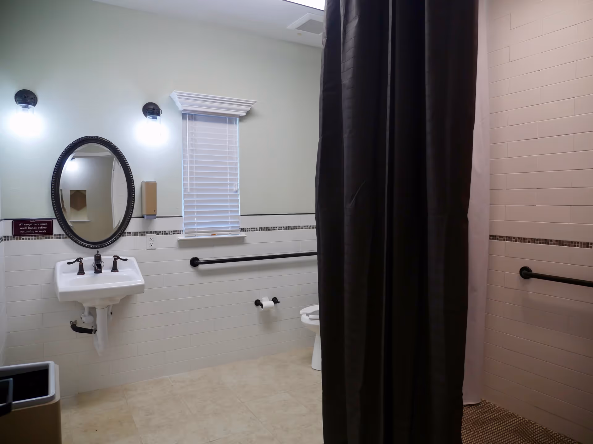 A clean bathroom with a white sink and oval mirror mounted on a light green wall. There are two wall lights above the mirror, a small window with blinds, a toilet partially visible behind a dark shower curtain, and grab bars on the walls for accessibility. The floor is tiled, and there is a trash can in the corner.